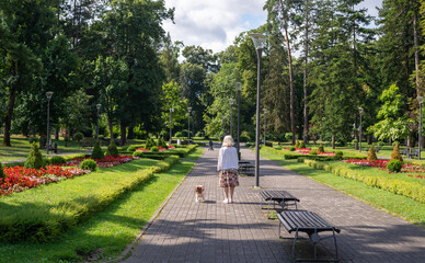 Woman and dog - Cavalier King Charles Spaniel - walking in a public park with lush flowers, plants,...