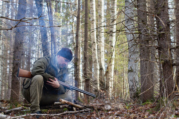 A hunter is squatting by a small campfire in the forest