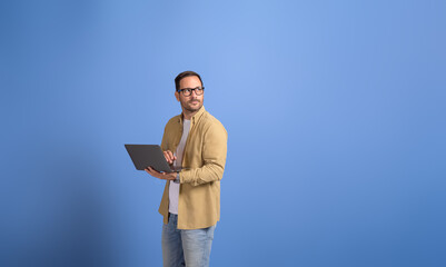 Serious young businessman holding laptop and looking away thoughtfully over isolated blue background
