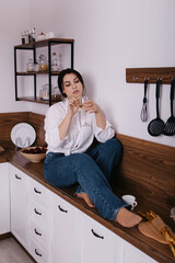 Young tipsy woman hold drink glass of champagne alcohol in kitchen sitting alone chilling relaxing on worktop wooden counter. Celebrating holiday, festive, celebration
