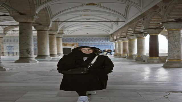 Smiling woman with glasses and headscarf standing in ancient ottoman architecture in istanbul.
