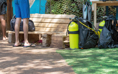 Aluminum Scuba dive cylinder gas air bottle tank for underwater activity. Two mans stand near a wooden bench surrounded by diving equipment. The equipment includes yellow oxygen tanks and black bags