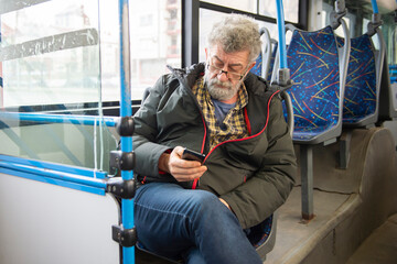 A man is sitting on a seat while riding a bus and having fun with his phone