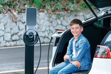 Little boy sitting on car trunk while recharging eco-friendly electric car from EV charging...