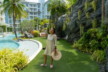 A lady stands in sunny weather on a green lawn between a swimming pool and an artificial rock with plants, holding a phone and a wide-brimmed hat in her hands and looking at the sky.