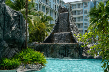 An artificial cascade of a waterfall whose water falls into a pool of blue water among tropical vegetation. Condominium at a resort in the tropics.