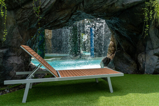An empty sun lounger standing in the shadow of a rock against the backdrop of an artificial waterfall with falling water in sunny weather at a tropical resort.