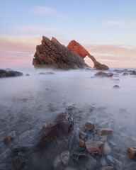 Landscape of natural sea arch and stones in the foreground covered with water. Famous rock formation on the Moray Coast, Scottish Highlands, Scotland. Bow Fiddle Rock at sunset, long exposure.