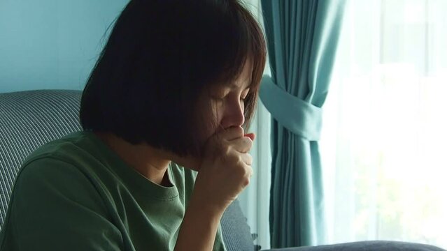 Asian woman sneezes or coughs with a tissue covering her mouth. Sick with a cold or allergic reaction to the weather on the sofa at home