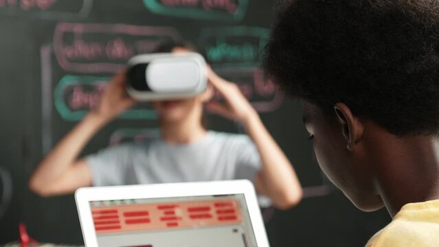 African boy programming system while caucasian girl enter metaverse while sitting at blackboard with engineering code written.high school girl wearing VR or headset in STEM technology. Edification