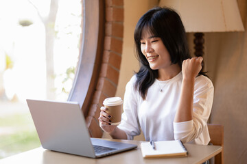 .Young Asian woman enjoying coffee while having a pleasant video call on a laptop in a coffee shop.