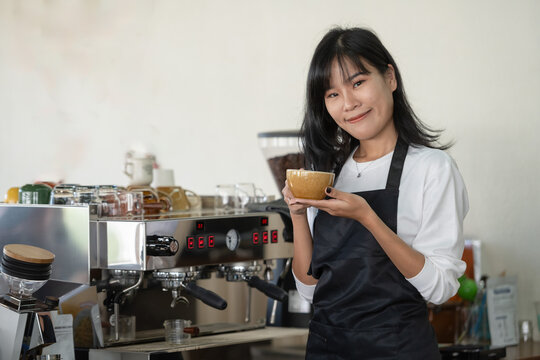 A smiling barista in a casual apron presenting a cup of coffee with professional espresso equipment in the background.