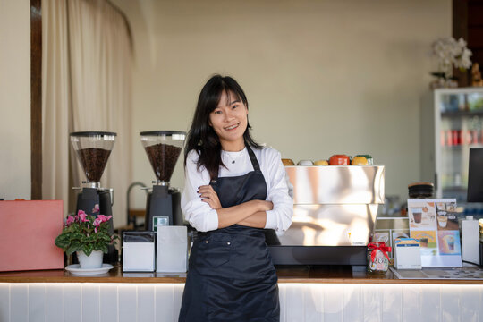 Young, cheerful female barista confidently standing at the coffee shop counter with arms crossed, surrounded by cafe equipment.