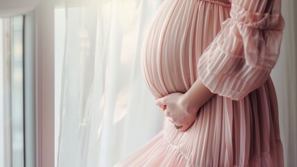 A pregnant woman in a pink dress stands in front of a window, looking out
