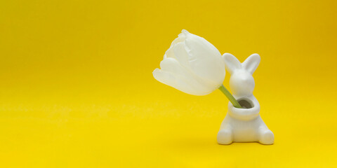 Ceramic figurine of Easter Bunny holds white tulip flower in hands on yellow background. Happy...
