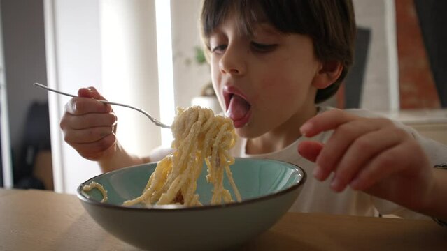 Child eating spaghetti, messy little boy spins pasta with fork at night mealtime