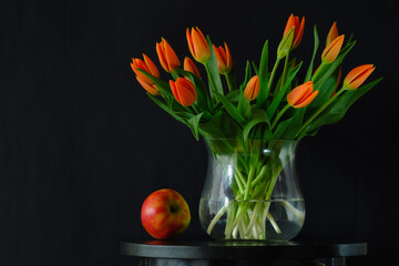 bouquet of orange tulips in a transparent round vase and an apple on a dark background.