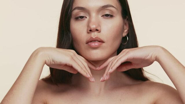 Close-up of a young woman in her twenties touching her face gently, portraying a beauty and skincare concept on a neutral background.