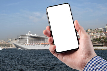 A man holds a smartphone in his hand. A large cruise ship stands on the pier with the city in the background.