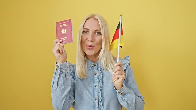 Joyful young caucasian blonde woman confidently holds german flag and passport, ready for a joyous european holiday, isolated on a cheery yellow background.