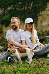 A young Caucasian couple is sitting in a green clearing among wildflowers and enjoying a peaceful walk with a Jack Russell terrier. People with small cute dog in the park are having fun outside.