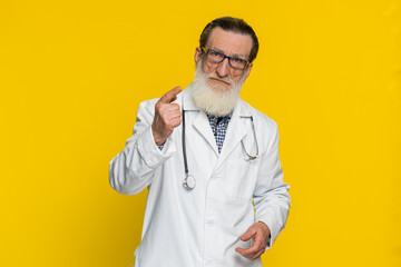 Senior doctor cardiologist man shakes finger and saying no, be careful, advice to avoid danger mistake, disapproval sign, disagree dislike quarrel. Apothecary pharmacy grandfather on yellow background