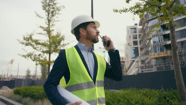 Caucasian foreman walking moving contractor civil engineer holding blueprint plan city building project guy male walk control construction builder man architect urban development talk walkie talkie