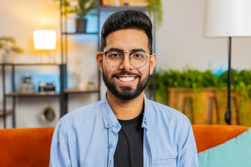 Portrait of happy Arabian man sitting on sofa looking at camera and smiling at home. Young Indian bearded guy wearing eyeglasses having attractive appearance relaxing thinking in living room.