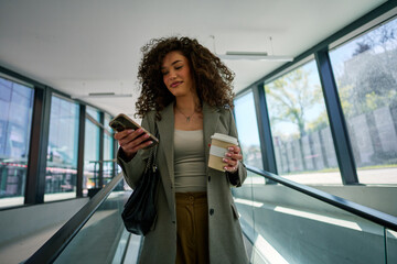 Joyful Caucasian woman with curly locks scrolling through her phone at the airport, coffee in hand...