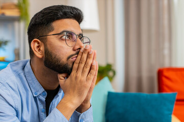 Tensed Indian man sits on sofa at home folding palms together pray ask for help thinks feel...