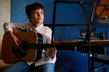 Hispanic teenage boy in white casual shirt, plucking strings while strumming guitar, playing...