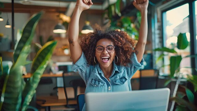 Excited young woman celebrating success at a laptop in a modern office. Indoor plant-filled workspace concept 
