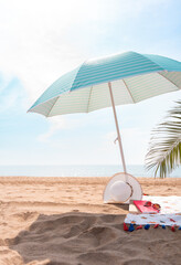 Beautiful summer morning with open turquoise umbrella, hat, sunglasses, book and towel on a beautiful lonely beach with a palm leaf on the right side.