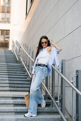 Young woman in a blue shirt and jeans stands on a set of stairs. She is wearing sunglasses and a white shirt and holding a handbag