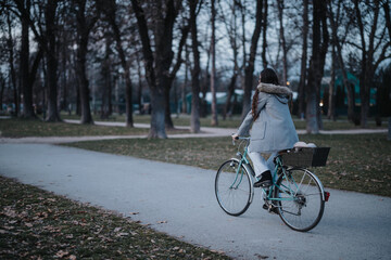 Back view of a young woman on a bicycle in a park, with trees and a footpath during the early evening.