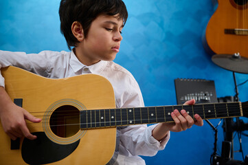 Adorable Caucasian teenager boy playing acoustic guitar in music studio. Electric guitar and drums...