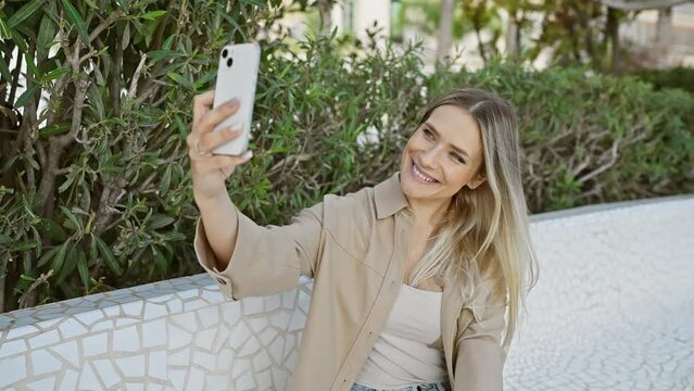 Cool, confident blonde young woman joyfully making a selfie with her smartphone, enjoying the city park's sunlit, green outdoors.