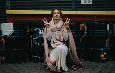 A self-assured entrepreneur shows a peace sign while seated confidently among whiskey barrels in an urban environment.