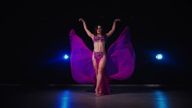 Woman in purple dress gracefully dancing belly dance in dimly lit room. Slow motion shooting. Elegantly dressed woman moving to music and dancing oriental dances in dark setting. Female dancer wearing