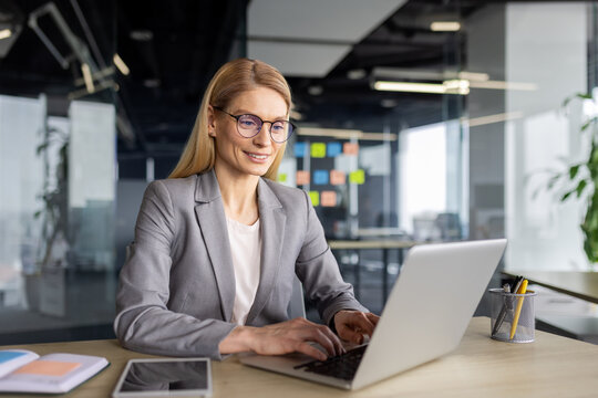 A cheerful mature businesswoman with glasses focused on her work, typing on a laptop in a modern office environment.