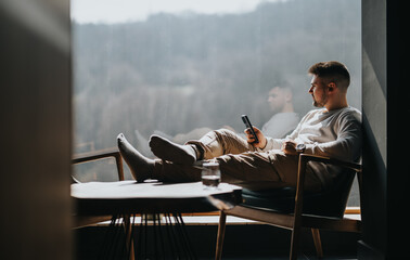 A young adult enjoys a peaceful moment in a brightly lit room, browsing his smart phone while...