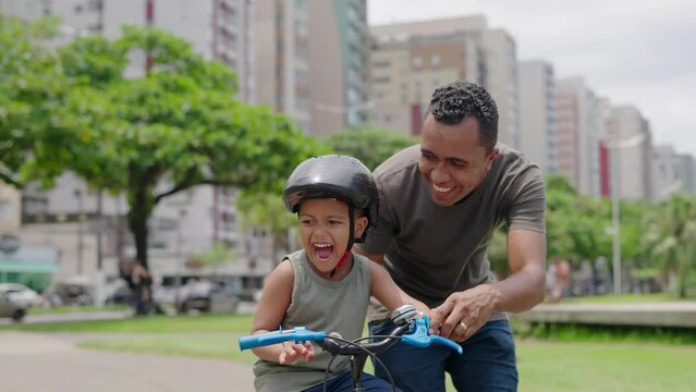 Happy latin father helping smiling boy to ride bike on cityu park. Happy latin child and young dad riding bike. Smiling daddy teaching son to ride a balance bicycle.
