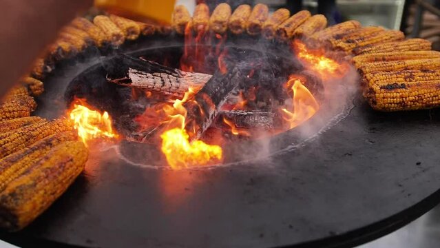 Burnt golden corncobs are cooked with smoke and flame on a grill or barbecue. Close-up of corn vegetables being cooked on a round metal grill. Picnic or party in the countryside. Summer weekend with h