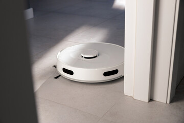 A white robot vacuum cleaner cleans grey tiles floors by the white door. collects dust and washes...