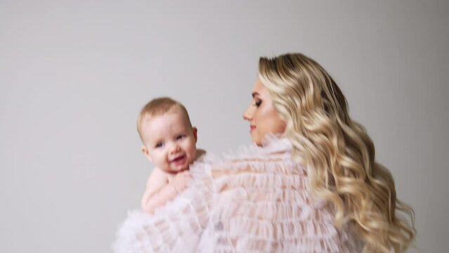 Caucasian woman with long wavy blonde hair turning around with baby in hands. Mother waving her cute child. White backdrop.