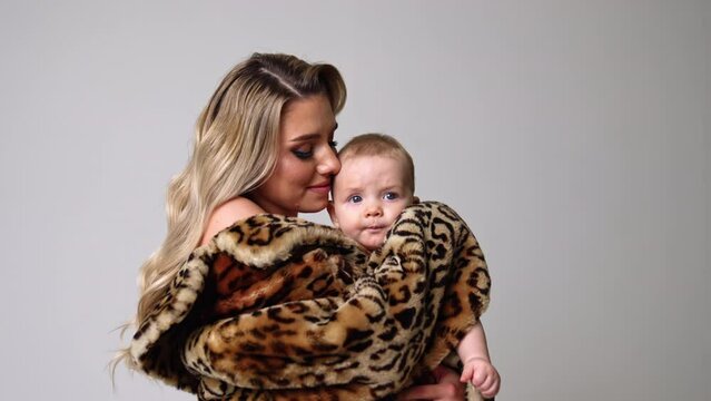Serious little infant baby in mom's hands. Woman wearing fur coat holds a kid with no clothes on. White backdrop.