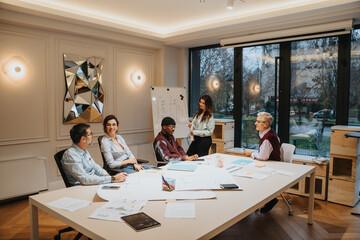 A multigenerational group of professionals engage in a lively brainstorming session in a well-lit office meeting room. They are focused on the presentation while sharing ideas and concepts.