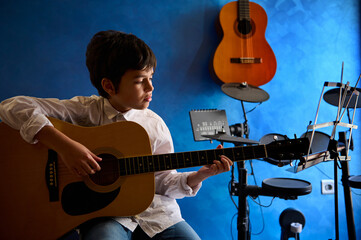 Adolescent boy strumming guitar in the modern music studio. Electric guitar hanging on a blue wall...