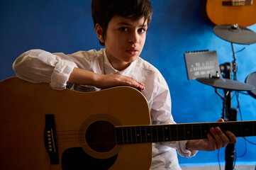 Handsome teenage boy playing guitar in the modern music studio, looking confidently at camera,...