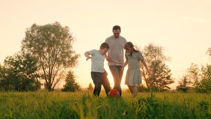 Naklejka na ściany i meble Family team, sports games. Children son daughter play football with father on lawn. Boy girl parent run after red ball in park. Active happy family, child dad having fun playing ball on green grass.